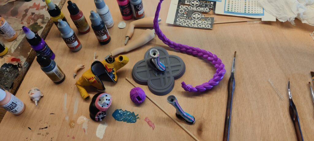 IMG_20260704_182048 ‣ Atelier 34 Craft table with bottles of acrylic paint, brushes, and doll parts scattered around; a purple braided piece and gray base in the center.