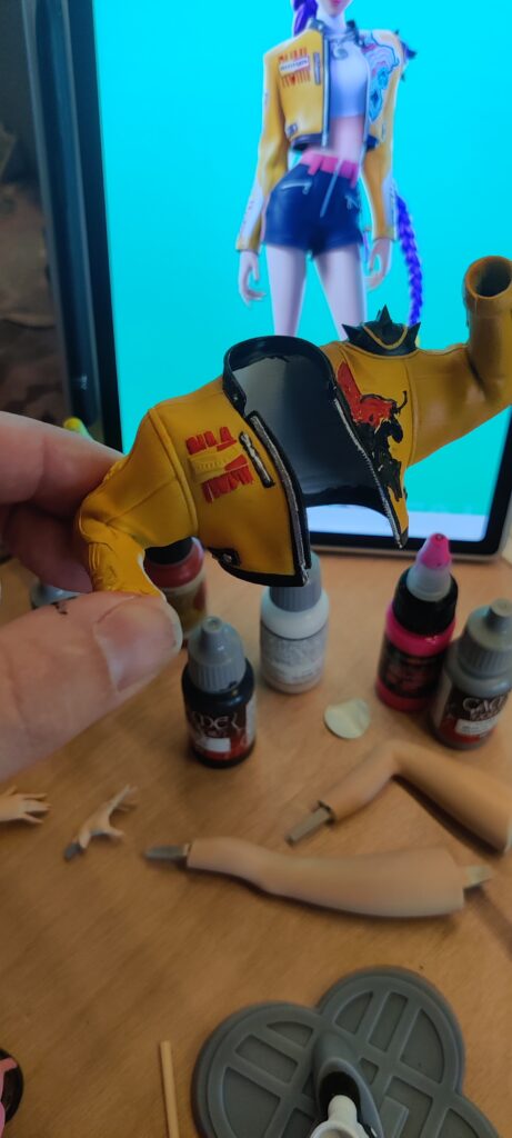 IMG_20260704_182138 ‣ Atelier 34 Close-up of a yellow toy jacket being held, with an open zipper, on a crafting desk with paints nearby.