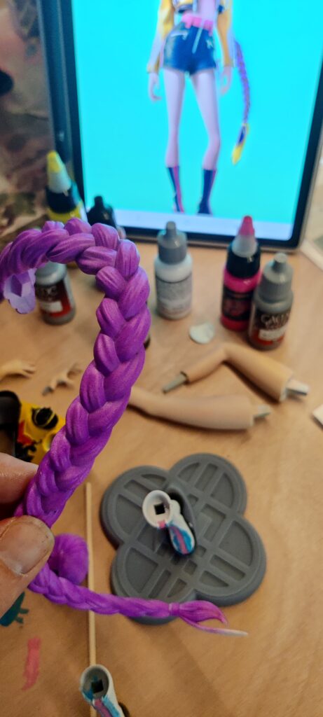 IMG_20260704_182255 ‣ Atelier 34 Purple braided bracelet held up in front of a crafting desk with paints, tools, and a mannequin figure on a screen in the background.