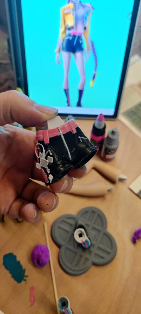 IMG_20260704_182309 ‣ Atelier 34 Close-up of a hand holding a small black doll shoe with pink trim and white patches at a craft station; a fashion image on screen in the background.