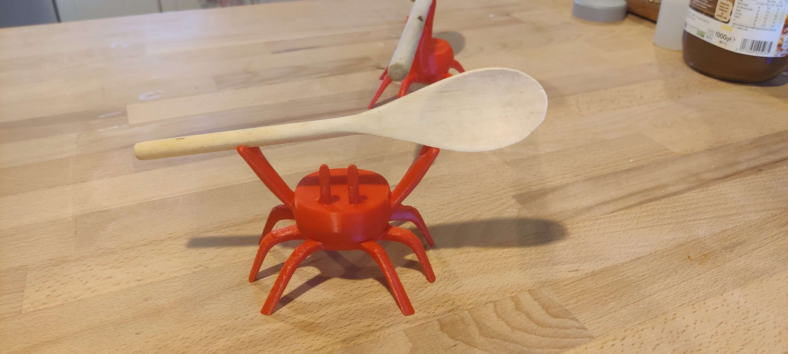 Crabe porte-cuillère en PLA – vue principale ‣ Atelier 34 Crabe rouge orangé imprimé en PLA tenant une cuillère en bois sur ses pinces