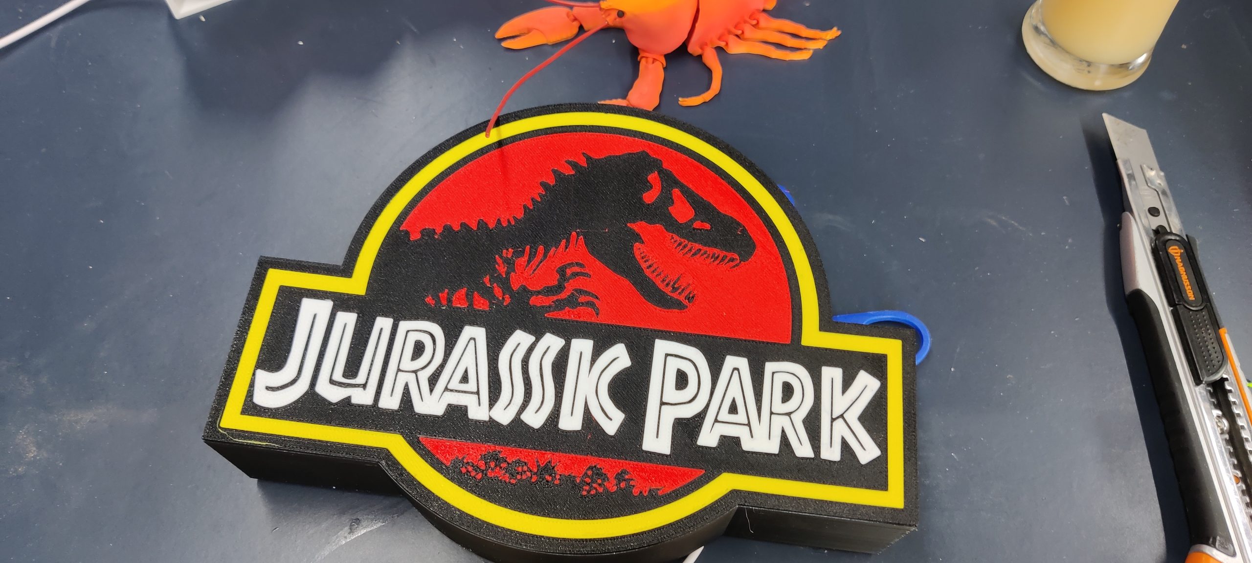 Lightbox Jurassic Park – Vue éteinte ‣ Atelier 34 Lightbox Jurassic Park éteinte, avec silhouette de T-Rex noire, fond rouge, contour jaune et lettrage blanc.